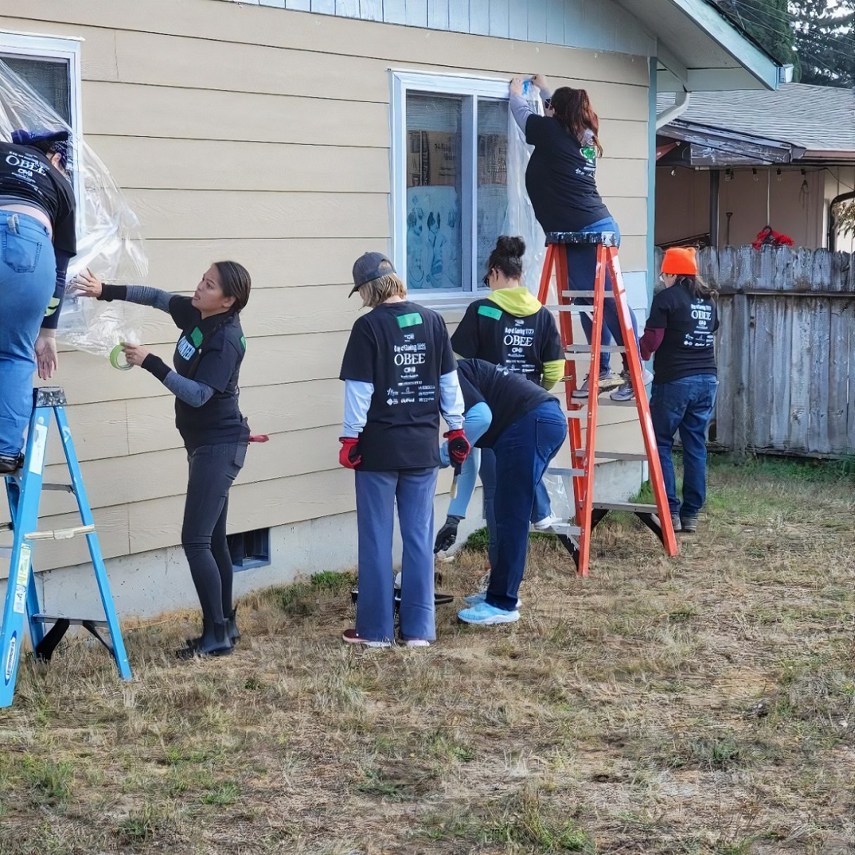 group of people in black tshirts prep the outside of a house for painting. Two are on ladders tapping plastic coverings on windows