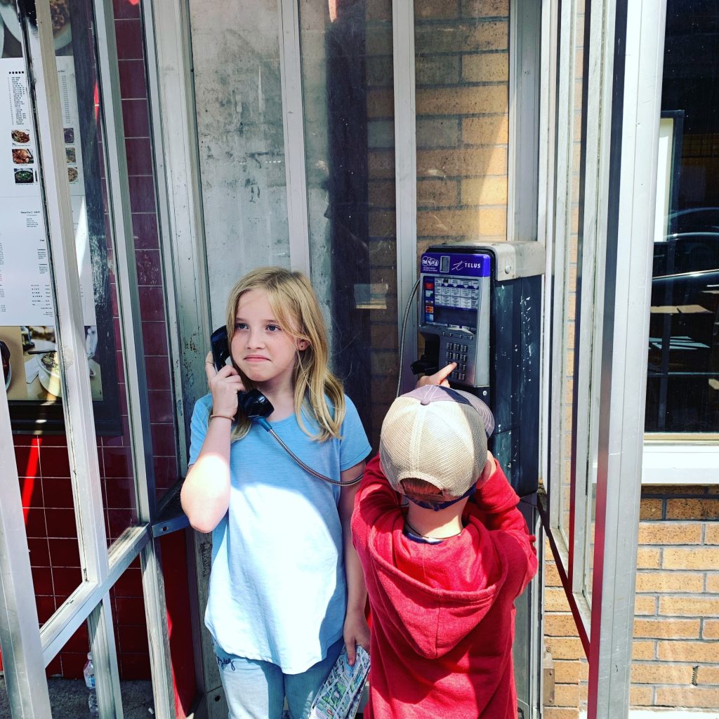 Two kids play in an old pay phone box. One is holding the receiver and the other is punching numbers