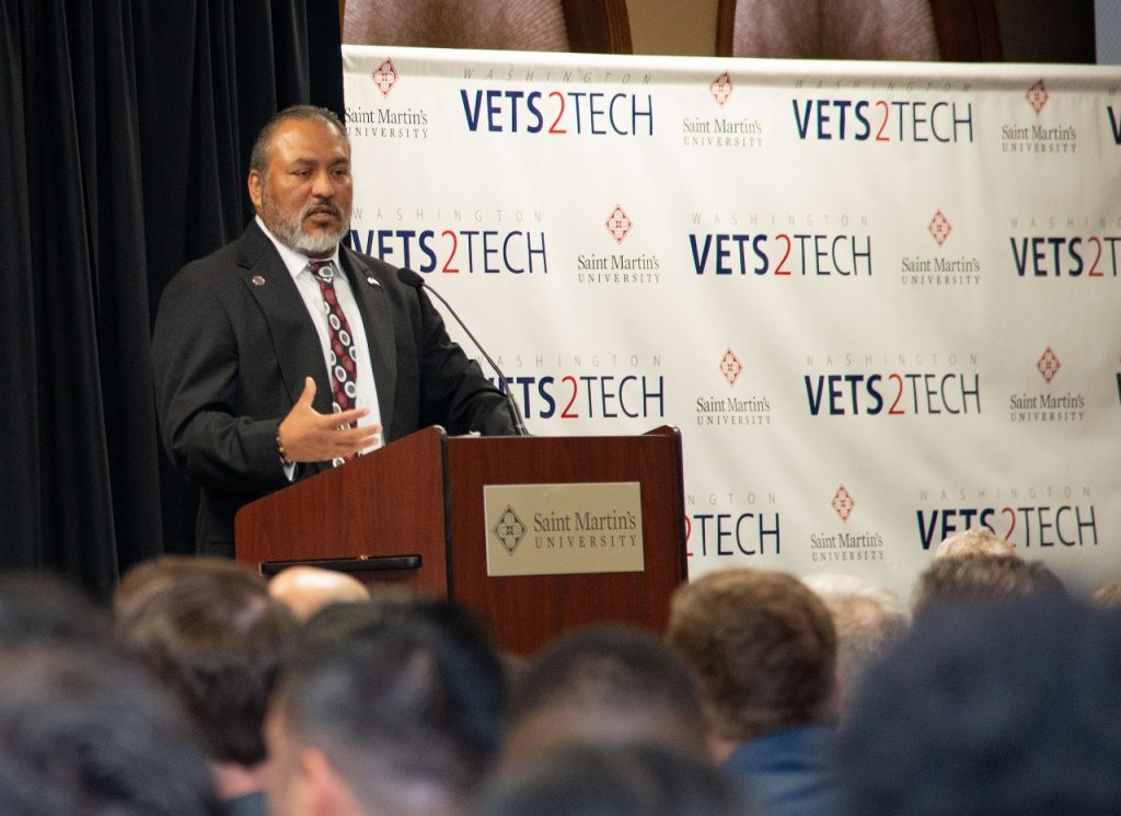 man in a suit stands at a podium with a step and repeat that says 'Vets2tech' behind him.