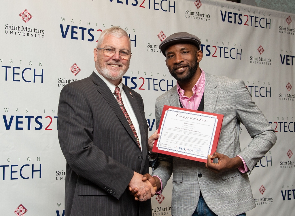 man in a suit shakes hands with a man holding a certificate with a step and repeat that says 'Vets2tech' behind them.