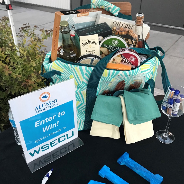 A tote bag full of wine, cheese, crackers, napkins and more on a black table with a wine glass and a sign that says, 'Enter to Win! WSECU'