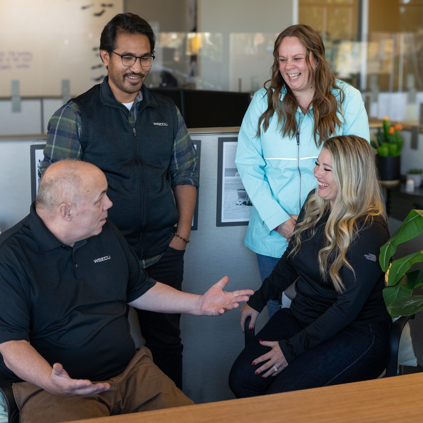 four people, two standing and two sitting, talk together in a bank cubicle