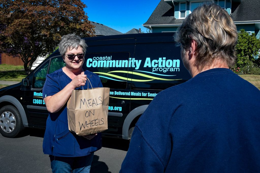 two people, one holding a brown paper bag, stand in front of a Meal on Wheels van.