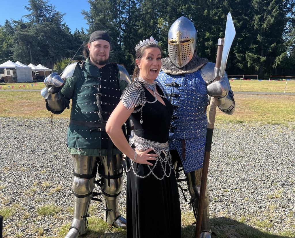 Linda Ciampi in a back skirt and halter top, wearing chainmail skirt and shoulder armor standing between two men dressed as knights
