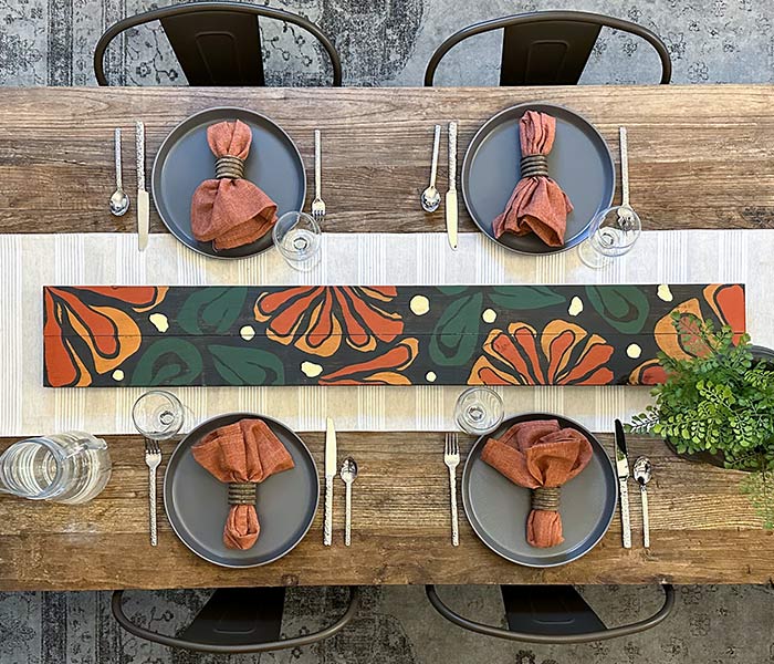 view of a table with a setting for four including napkins, and a long wooden painting running down the middle on top of a cream runner. The wooden painting has orange and green designs.