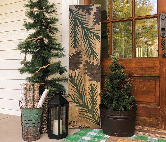 a wooden sign with pineconesa nd pine branches painted on it that says, 'The Bernettes' leans against a house with fake fir trees next to it and birch logs in a tin can.