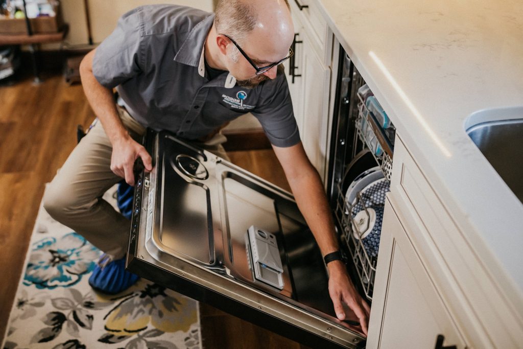a man kneels in front of a dishwasher, opening it up with one hand and using the other hand to feel along the door hinge