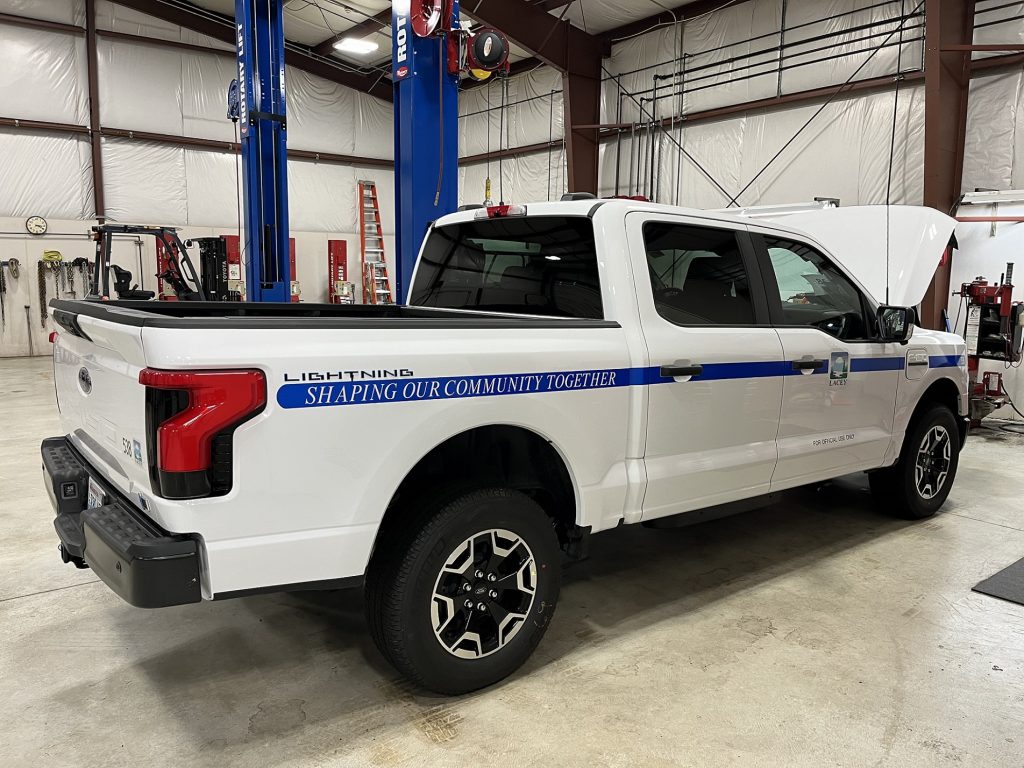 White electric pickup in a large garage with its hood up, the words, 'lightening, shaping our community together' are in blue on the side of the truck