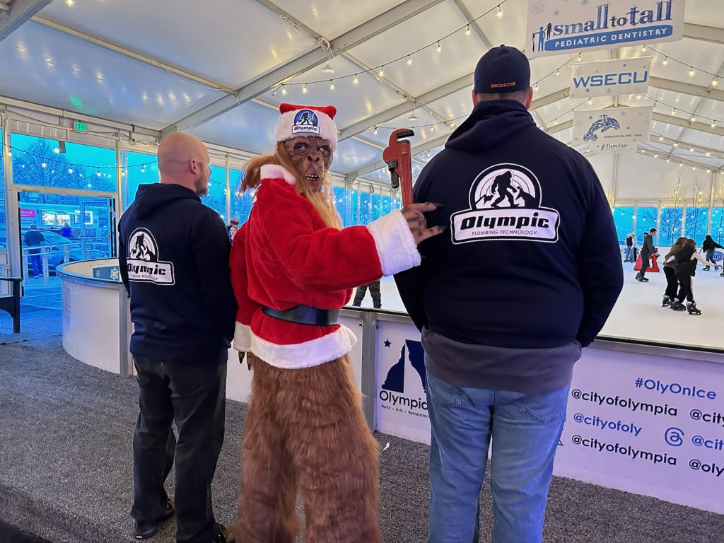 Two men in Olympic Plumbing Technology sweatshirts face the ice rink with a person dressed as bigfoot in a santa hat and coat stands between them and looks back over his own shoulder