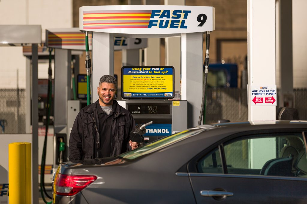A person stands by a grey car getting gas at Fast Fuel