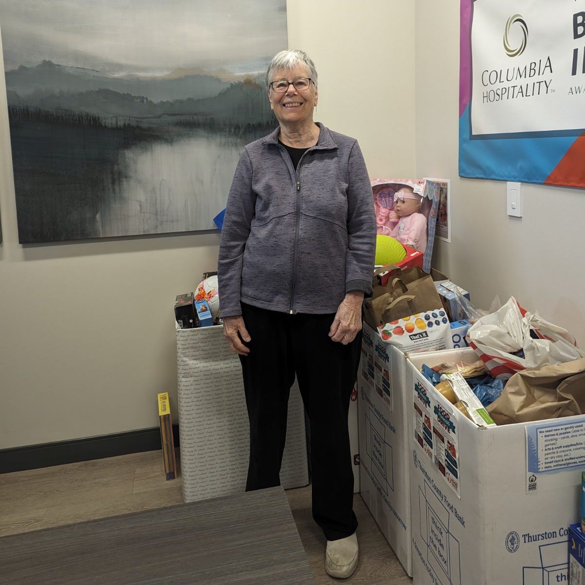 Kathie Deviny standing by larges boxes full of donated item. A landscape painting of a lake and mountains is on the wall behind her