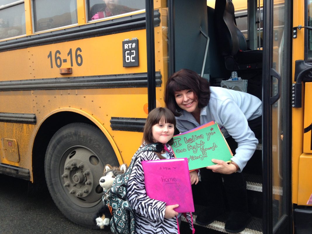 a student poses with her bus driver on the steps on the bus. the bus driver is holding a green piece of paper that says, 'I believe you can fight through anything'