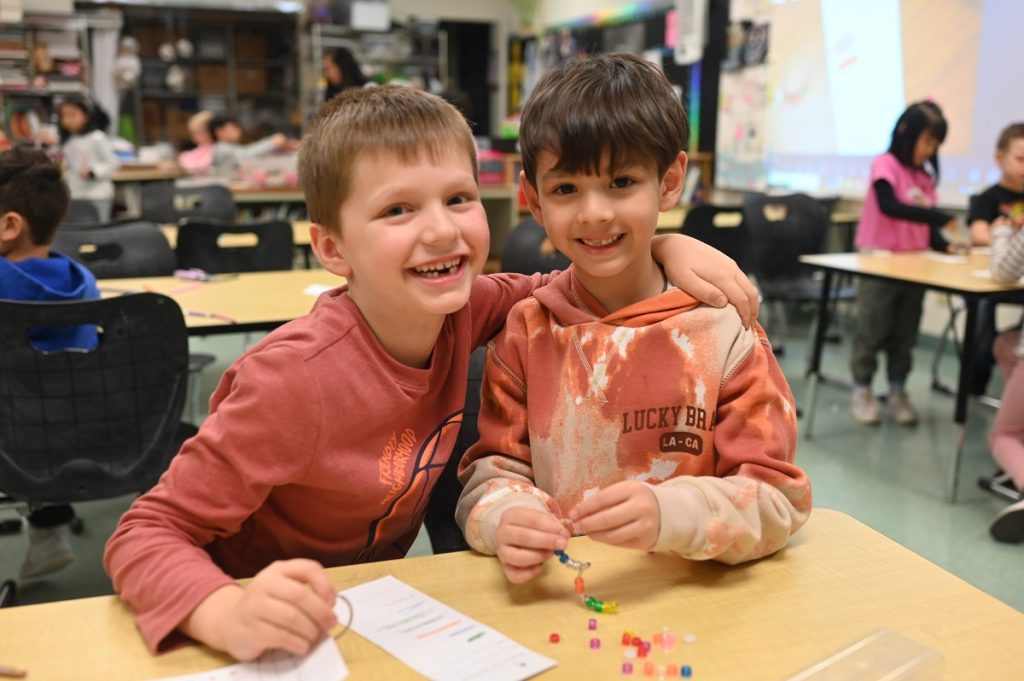 two young boys pose at a table, one with his arm around the other, both are smiing