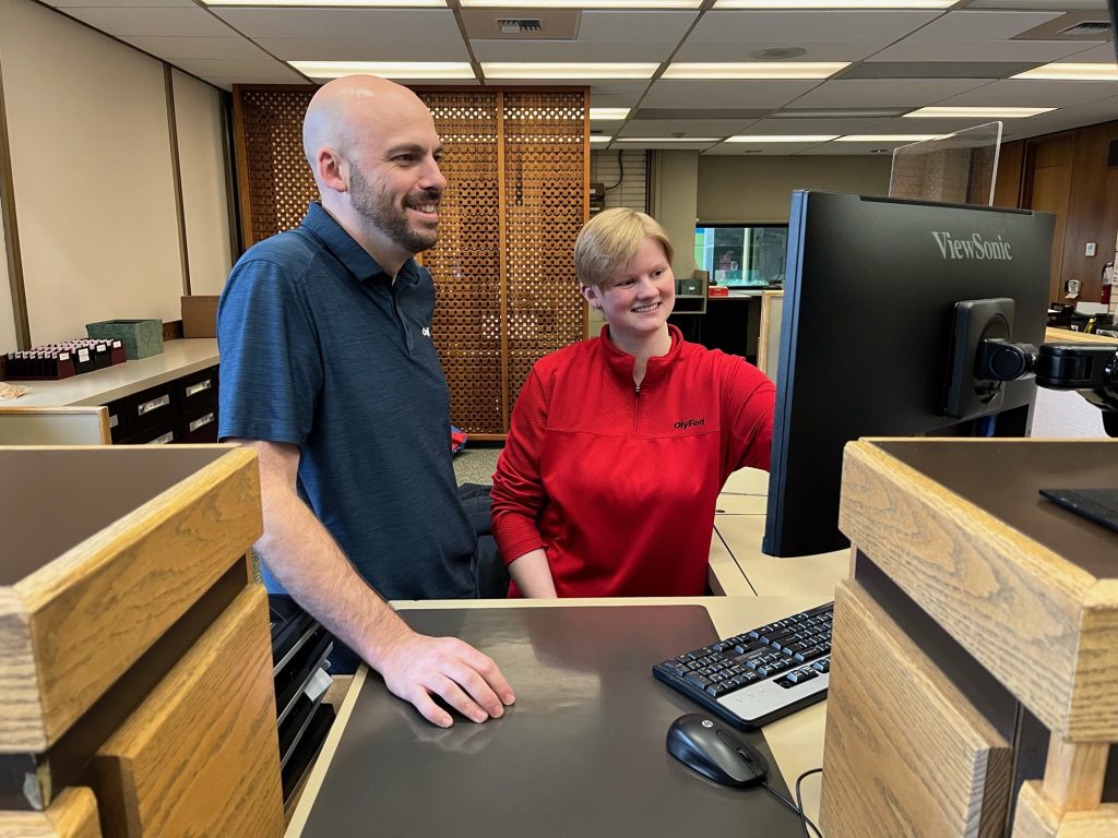 A man and a woman stand at a tall desk looking at a computer