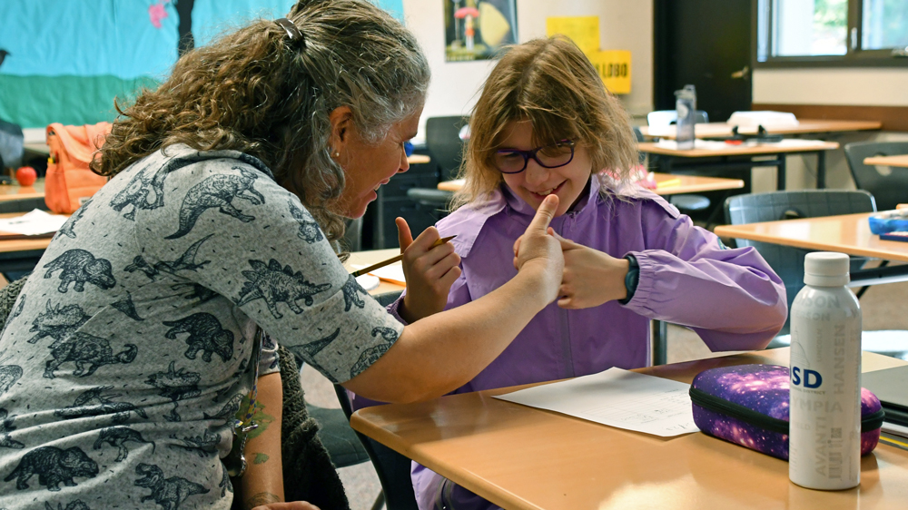 a teacher gives a young student a thumbs up while sitting next to her. The girl is also giving the thumbs up