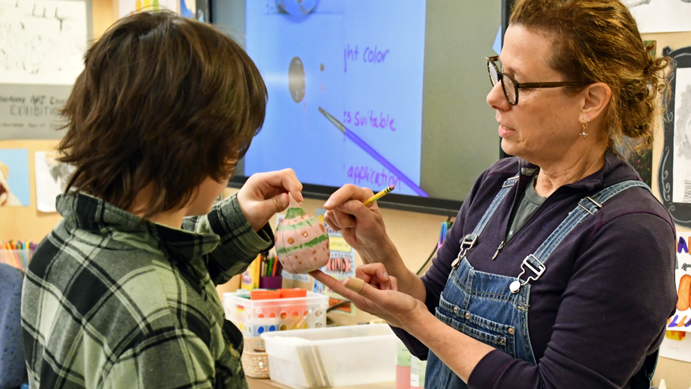 a teacher helping a middle schooler with a paper art project - the teacher is holding an egg shaped piece of art with pink and green watercolors on it
