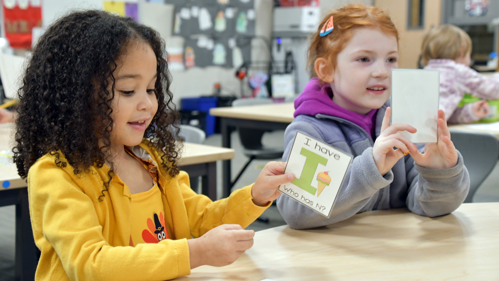 a young girl sitting in a classroom holds up a laminated flash card with the words 'I have 'I" who has N?' and an ice cream cone drawing next to the I.