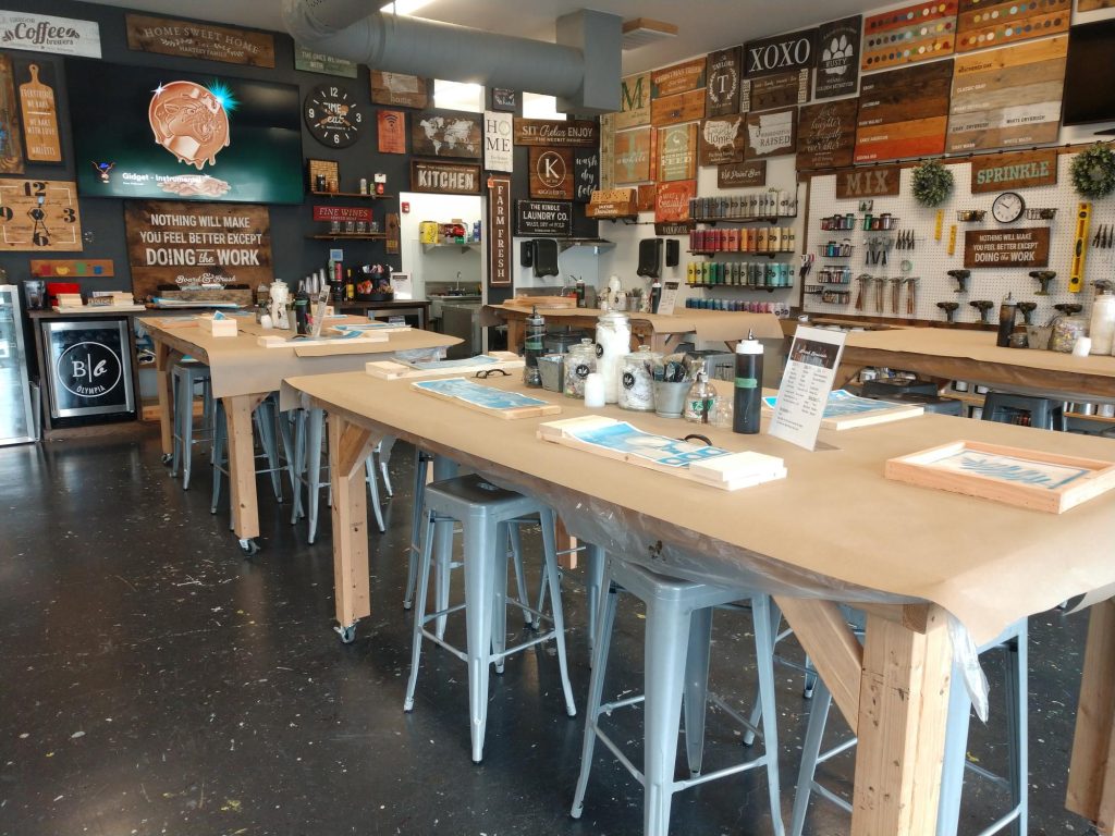 large wood tables with metal stools underneath are covered with butcher paper and have an assortment of art supplies on top. Wood signs hang all over the walls