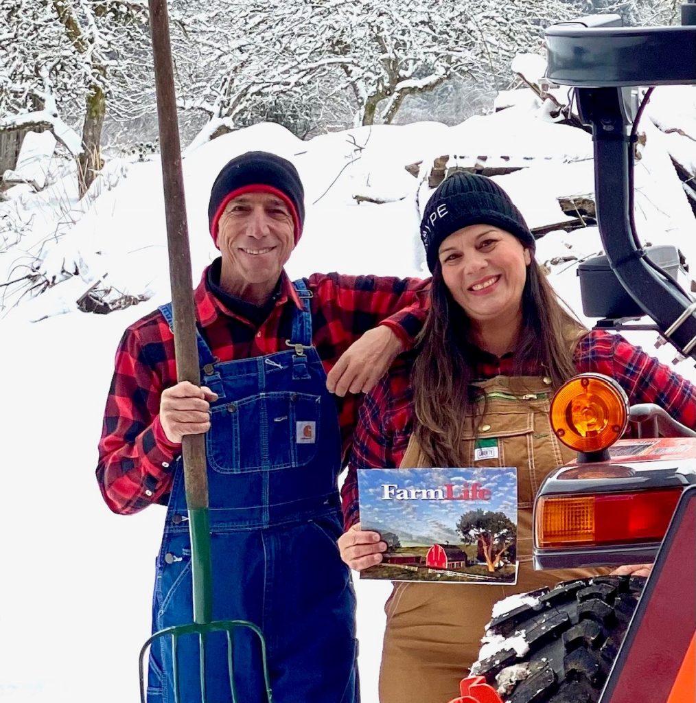 Mitch and Michelle Lewis in overalls in the snow, standing by a tractor. Mitch is holding a pitchfork and Michelle is holding a Farmlife calendar