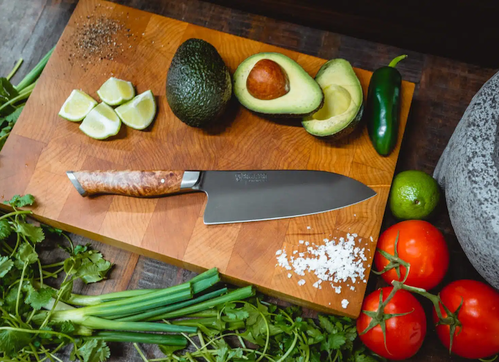 a STEELPORT knife on a wood cutting board with avocados cut in half and tomatoes and chives around it