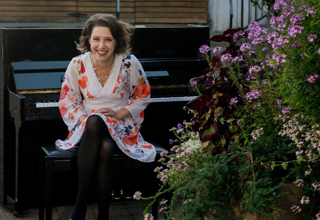 Sarah hagen in a white floral dress sitting on a piano bench with a piano behind her and flowers in front of her