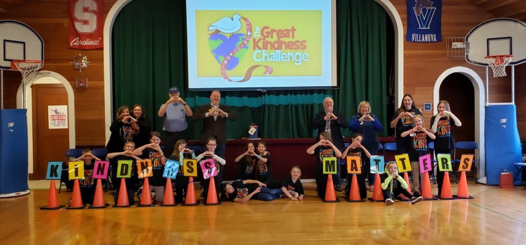 kids and staff in a Yelm school gym, holding their hands up to make a heart shape and orange cones with letters spelling out 'Kindness matters' in front of them. A screen behind them says 'The great Kindness Challenge' and has a heart-shaped Earth with dove sitting on it