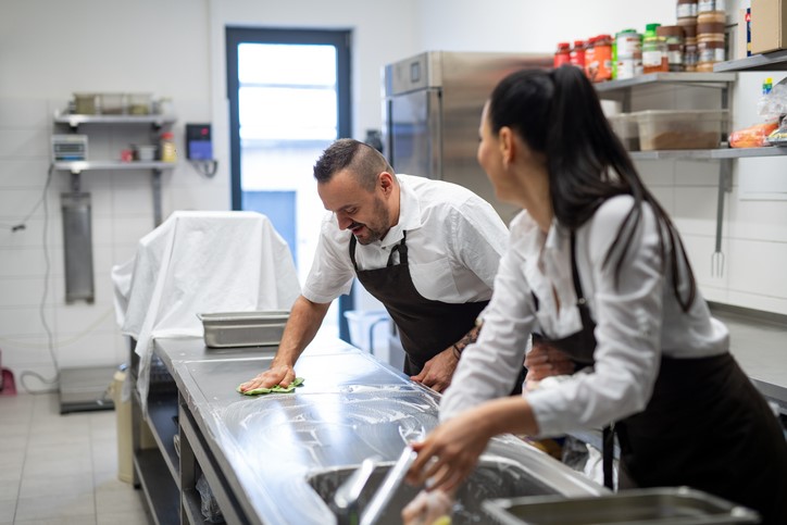 two people in black aprons clean a metal countertop in a commercial kitchen