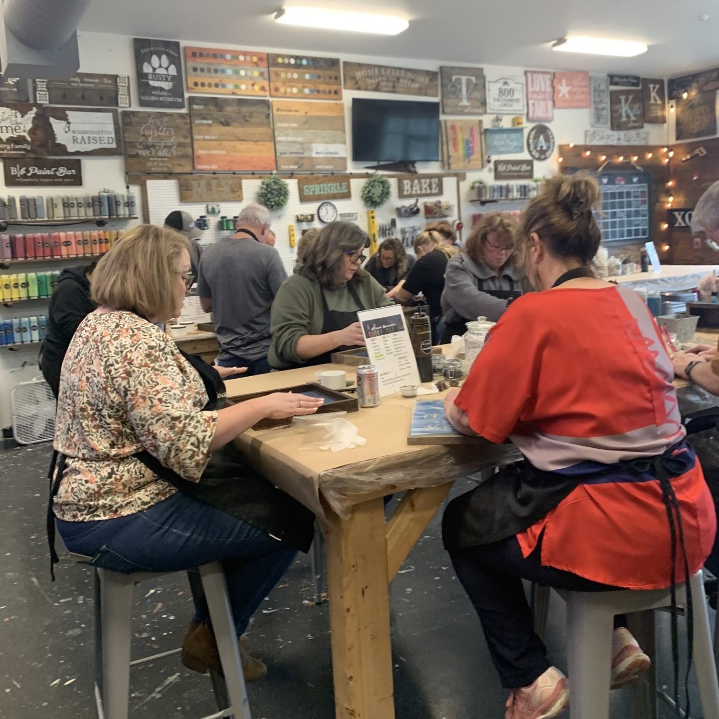 People in aaprons sitting on stools  at high wooden tables working on DIY Make and take projects at Board & Brush