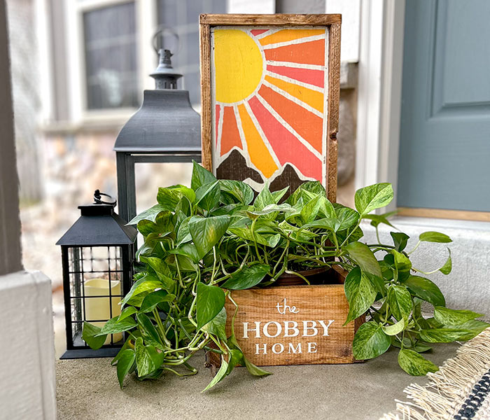 a wooden box that says "the hobby home" with fake ivy in it, a wooden sign painted with a sun design and two lanterns on a front porch.