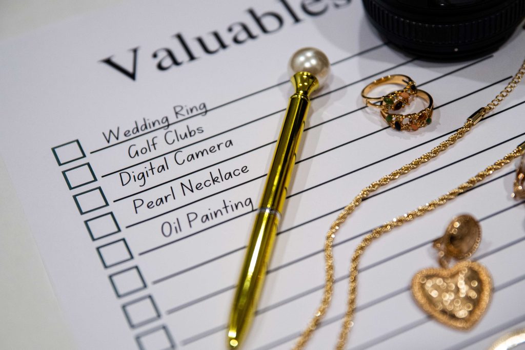 piece of paper that says valuables and has check boxes next to the words wedding ring, golf clubs, digital camera, pearl necklace and oil painting. A gold pen, gold chain, gold heart and gold rings lay on the paper.