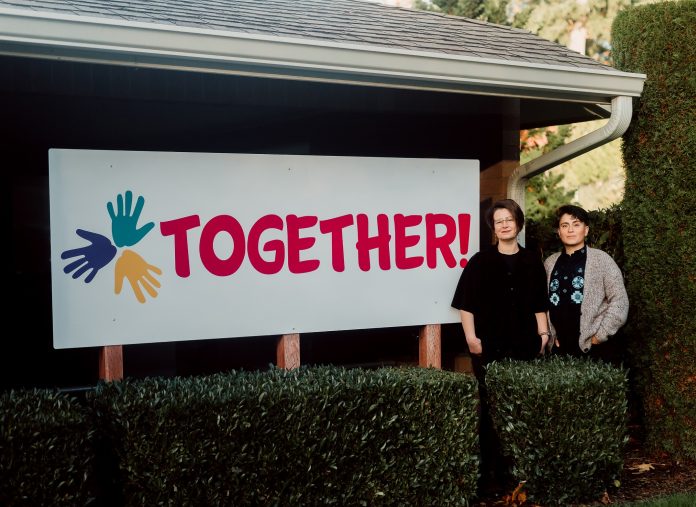 olympia-TOGETHER-co-directors-Kahlo-and-Sierra Sierra Abrams (left) and Kahlo C. Flores M. (right). standing by a white sign with three handprints on it and the word, 'TOGETHER!'