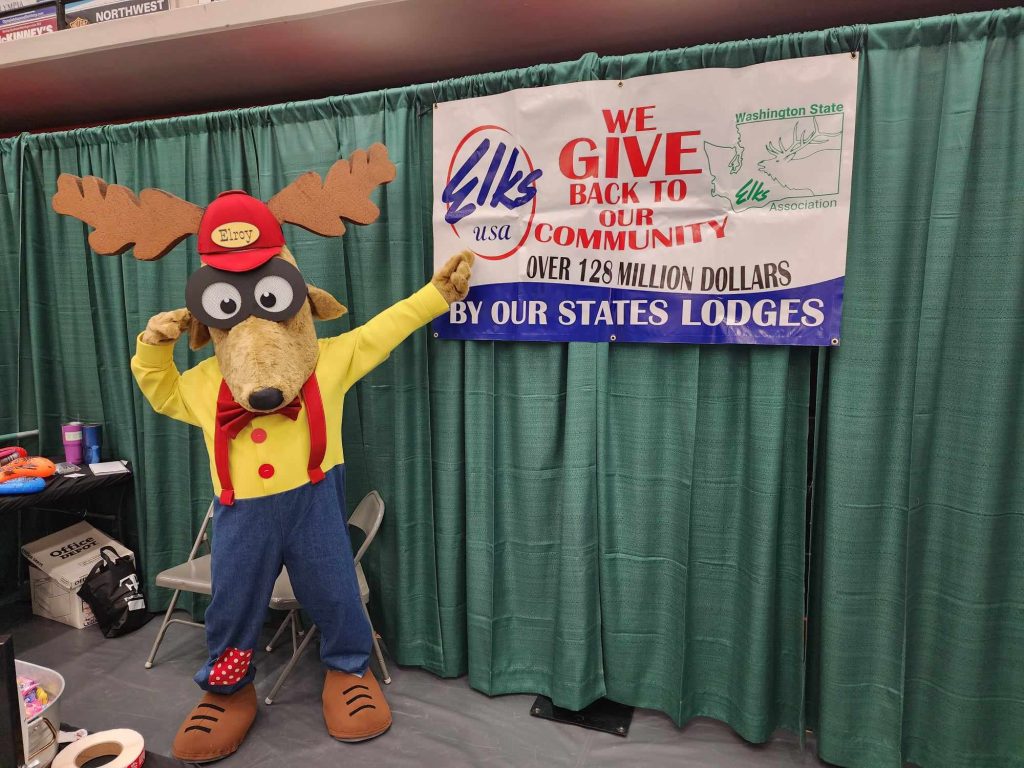 Elk Lodge mascot Elk pointing to a sign that says 'We give back to our community. Over 128 million dollars by our states lodges'