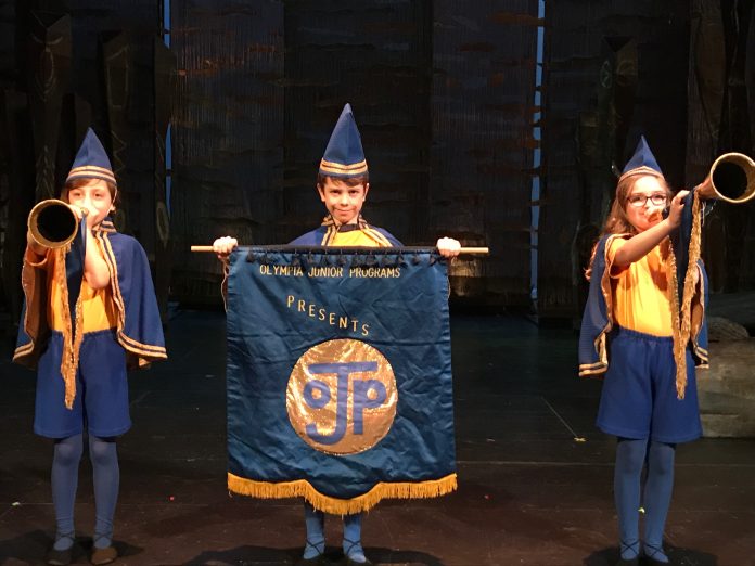 three little kids on stage, one holding a large banner, the other blowing on old-fashioned trumpets