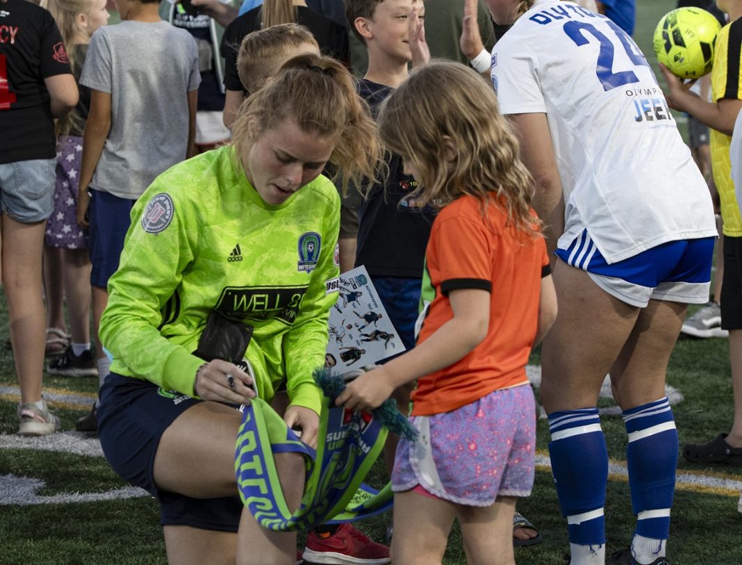 woman soccer player kneeling down to sign something for a young girl fan at a game, crowd in the background