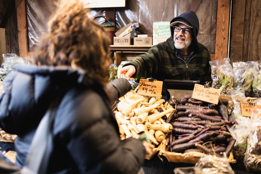 a woman buys vegetables from a man at a booth at the Olympia Farmers Market