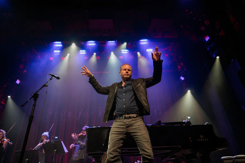 John Ondrasik with his hands up on stage, there is a string quartet behind him