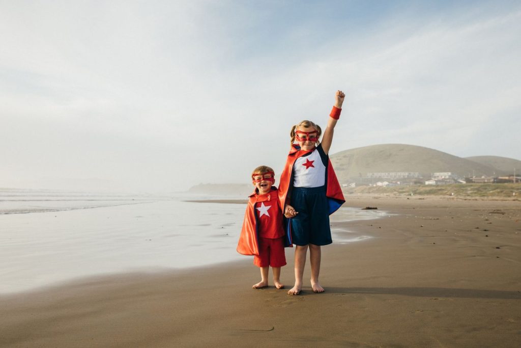 A young girl and a young boy dressed as superheroes stand barefoot on a California beach, the girl with her arm raised to the blue sky. They are siblings holding hands. The sister is ready to save the world, but her brother is still timid. She is a good example of confidence and has the strength to overcome all adversity. Image taken at Morro Bay, California.