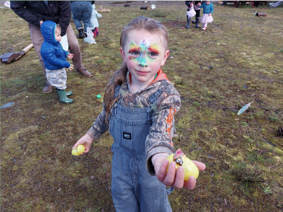 Little girl in overalls with her facepaintined holding up an Easter egg