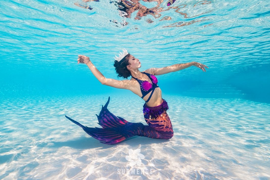 a woman in a mermaid tail knees on sand beneath the water