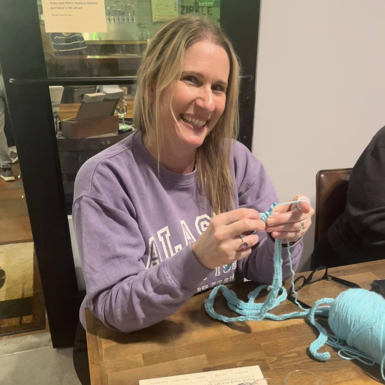 a woman smiles while crocheting at a table with other knitters and crocheters