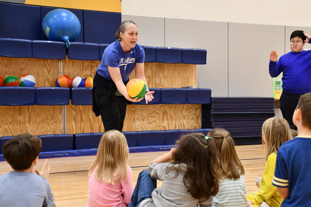 Kristen Draper showing kids sitting on a gym floor how to serve a volleyball underhand