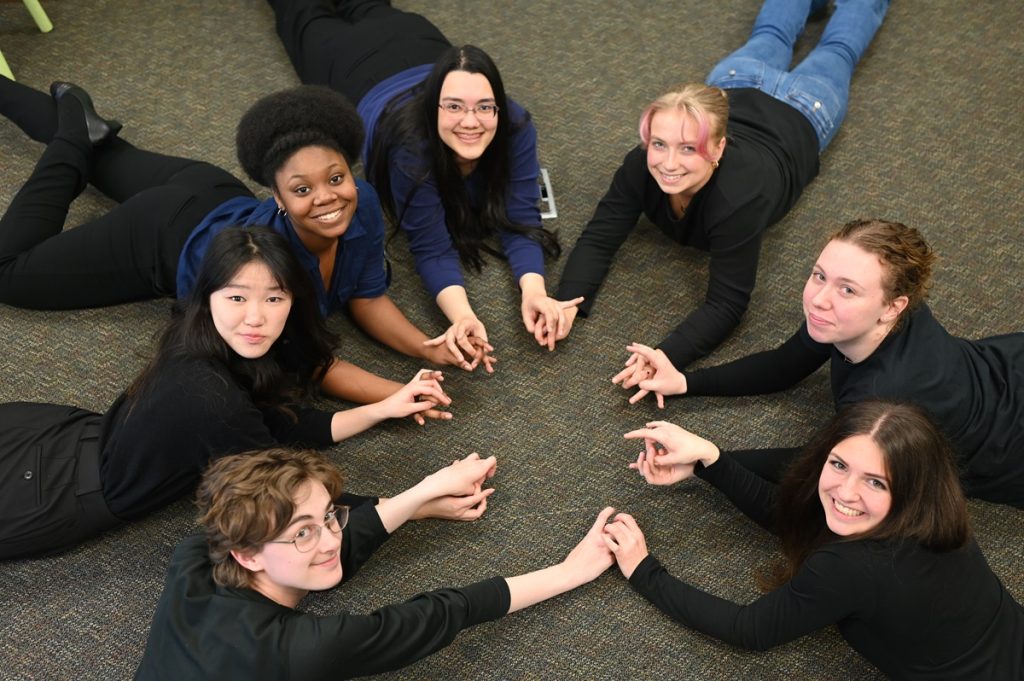 River Ridge High School ASL 3 students lying on the floor on their stomachs holding hands in a circle, smiling at the camera