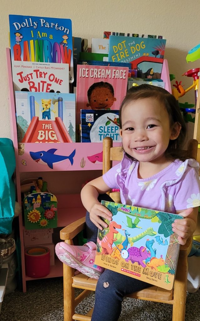 young girl in a chair with a book with a bunch of books in a shelf behind her