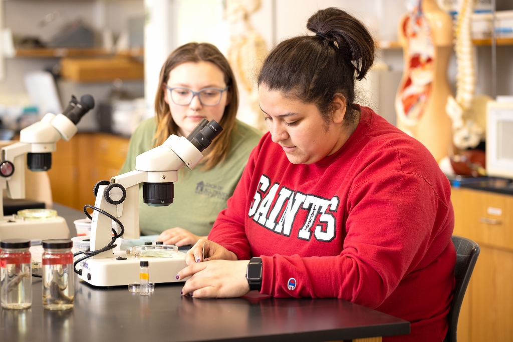two female students sit at a classroom table with microscopes in front of them