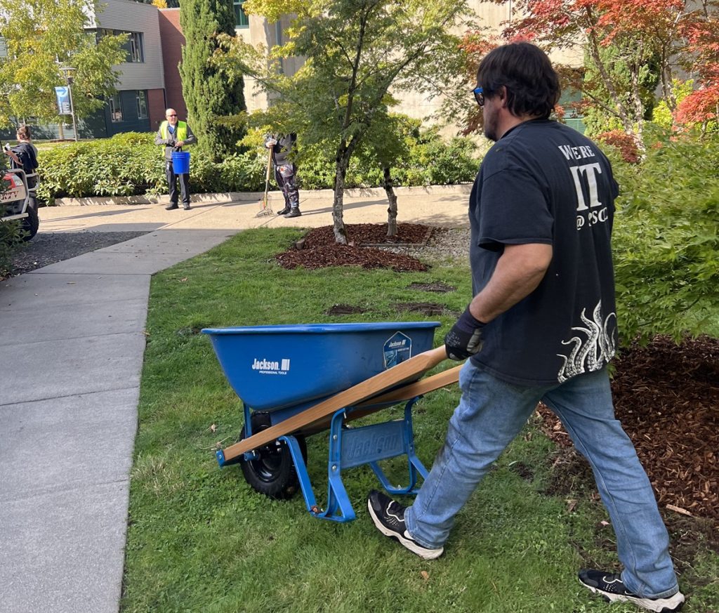 man wheeling a blue wheelbarrow over some grass, people working in the landscape are in the background.