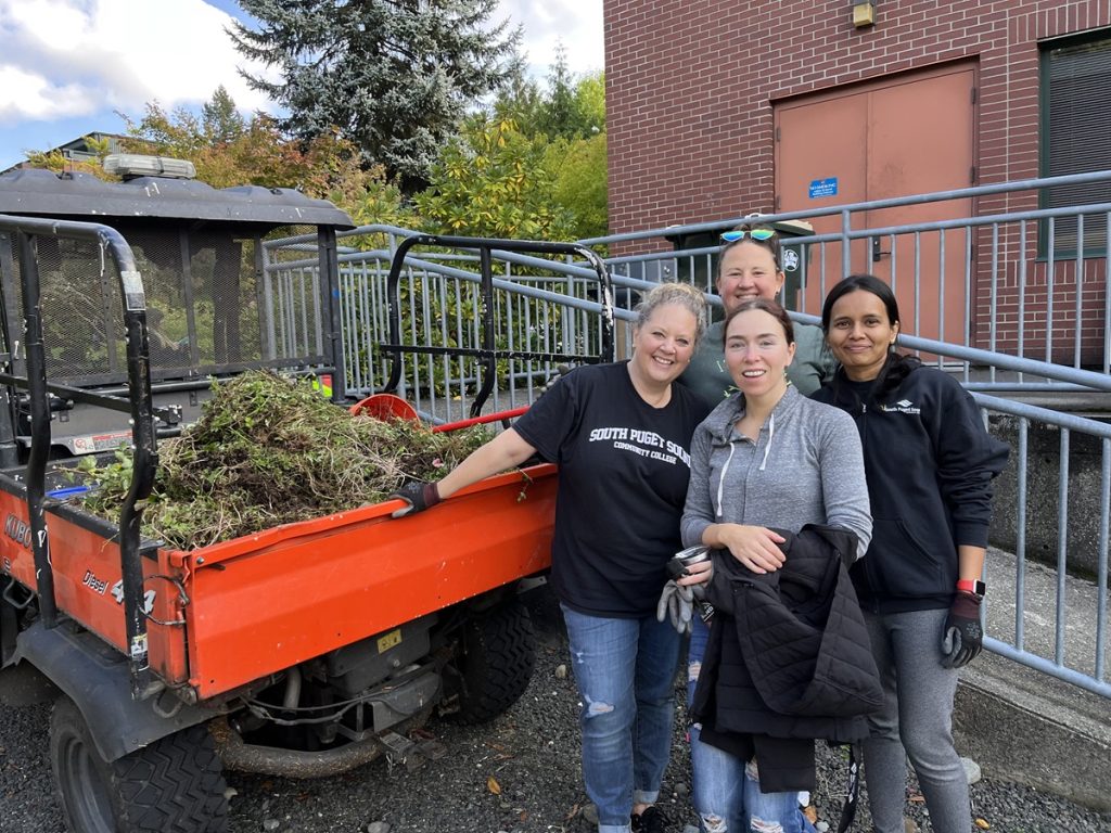 a group of four people stand by a large red container full of yard debris.