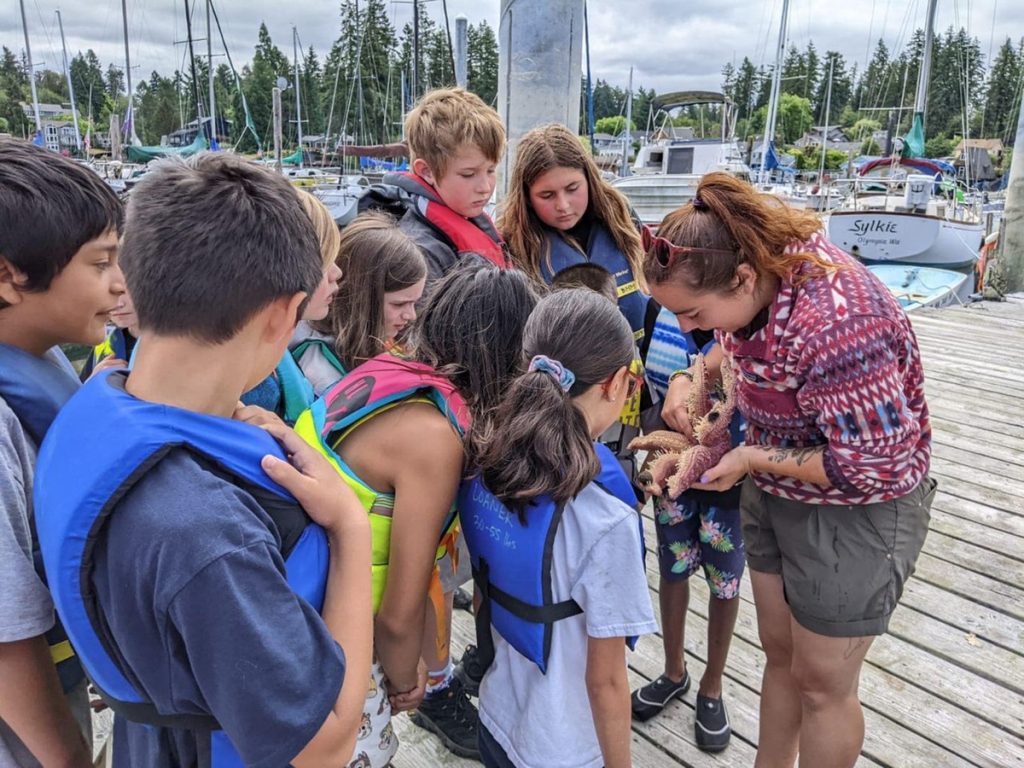kids with life vests on look at something a  Wa-Ya camp counselor is holding while standing on a dock