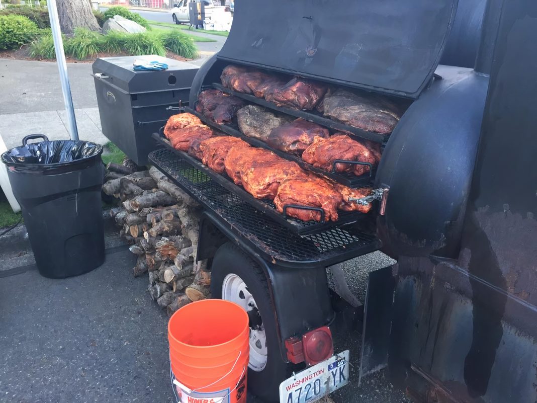 a huge black trailer grill full of meat cooking on it, firewood is stacked beneath it