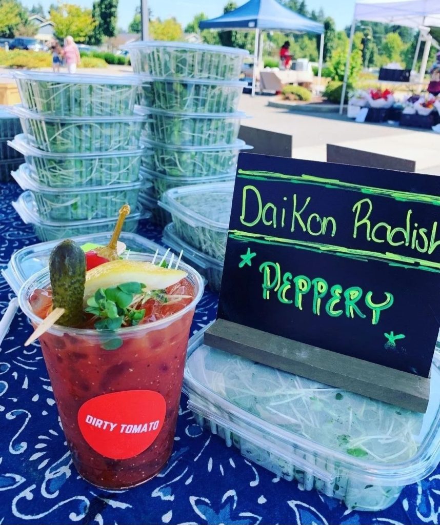 a red iced drink with a pickle, peper, lemon slice on top with a sticker that says "dirty tomato' sitting on a table next to a sign that says Daikon Radish, peppery.