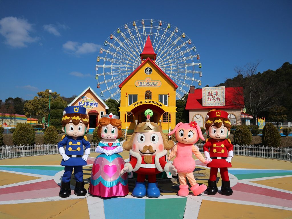 People in large animated costumes of a policeman, princess, kin, dog and soldier standing in front of a Yellow building that says 'toy kingdom' with a ferris wheel behind it